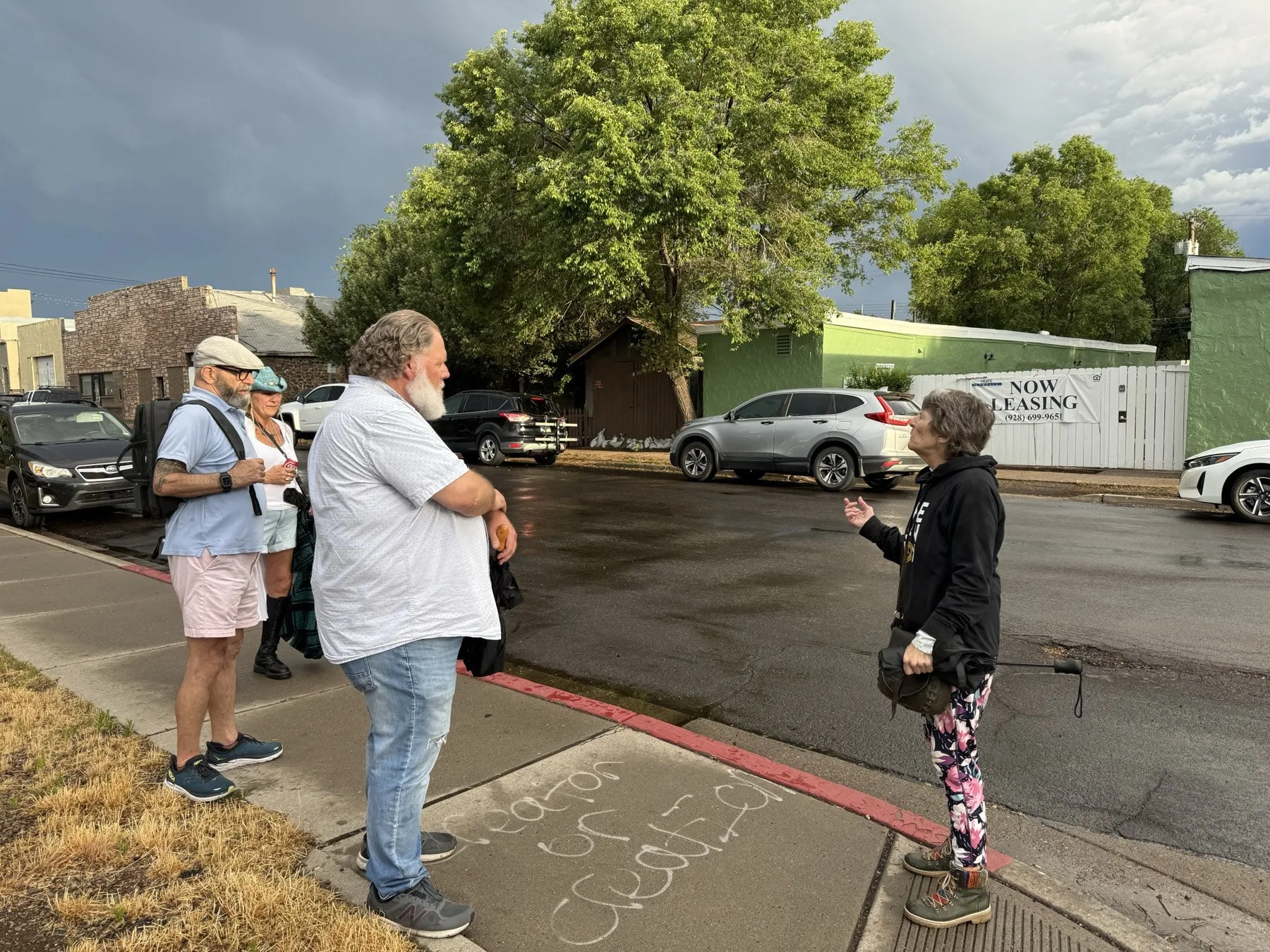 A Freaky Foot Tours guide talks with guests on a Southside Flagstaff sidewalk under a monsoon sky, old stone storefronts behind them